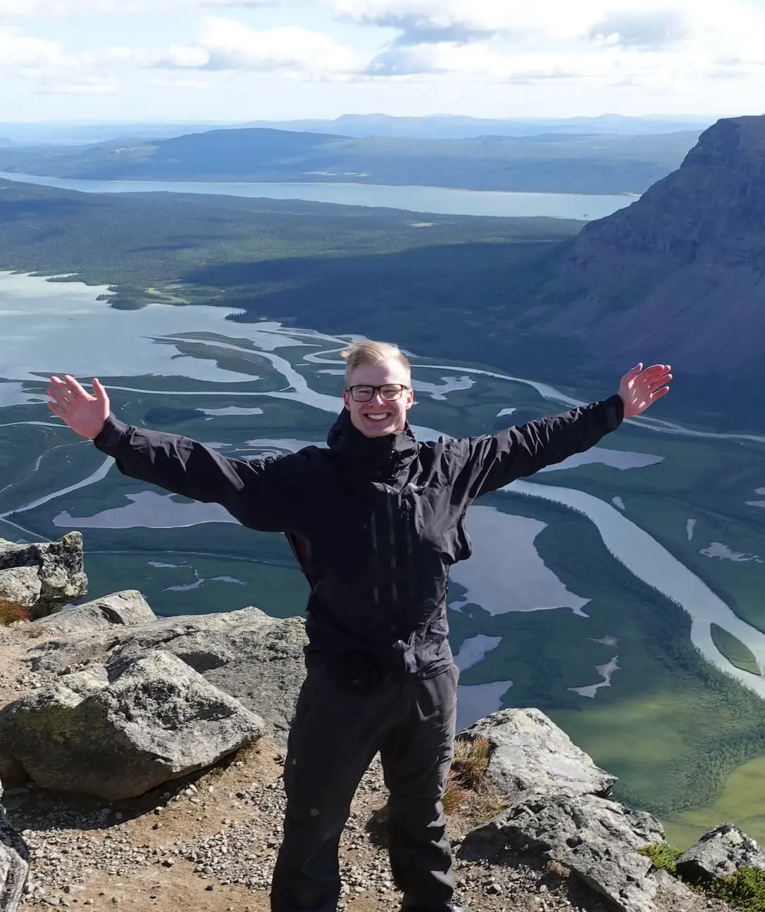 Ung man med blont kort hår och glasögon står på toppen av ett berg med ett delta i bakgrunden med flera vattendrag som slingrar sig genom ett grön landskap